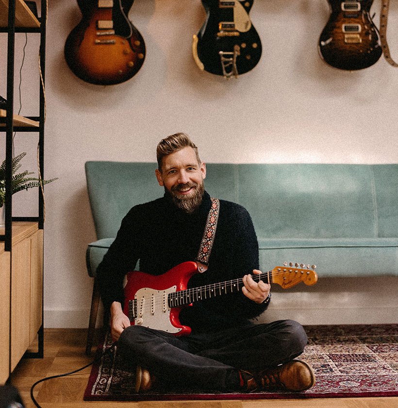 Paul Davids seated with a red Stratocaster in front of a wall of vintage guitars.